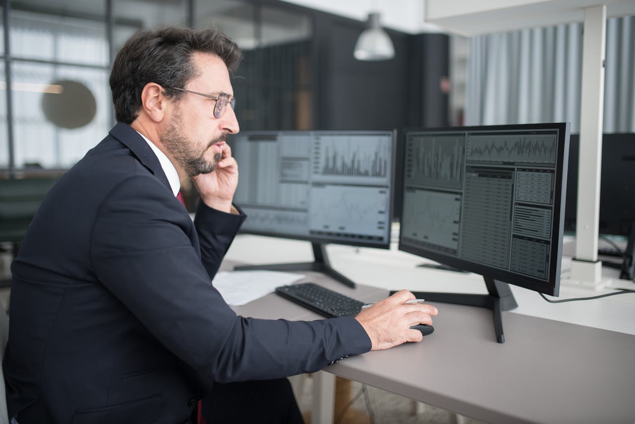 services-02 Businessman analyzing stock market data on dual monitors in a modern office setting.